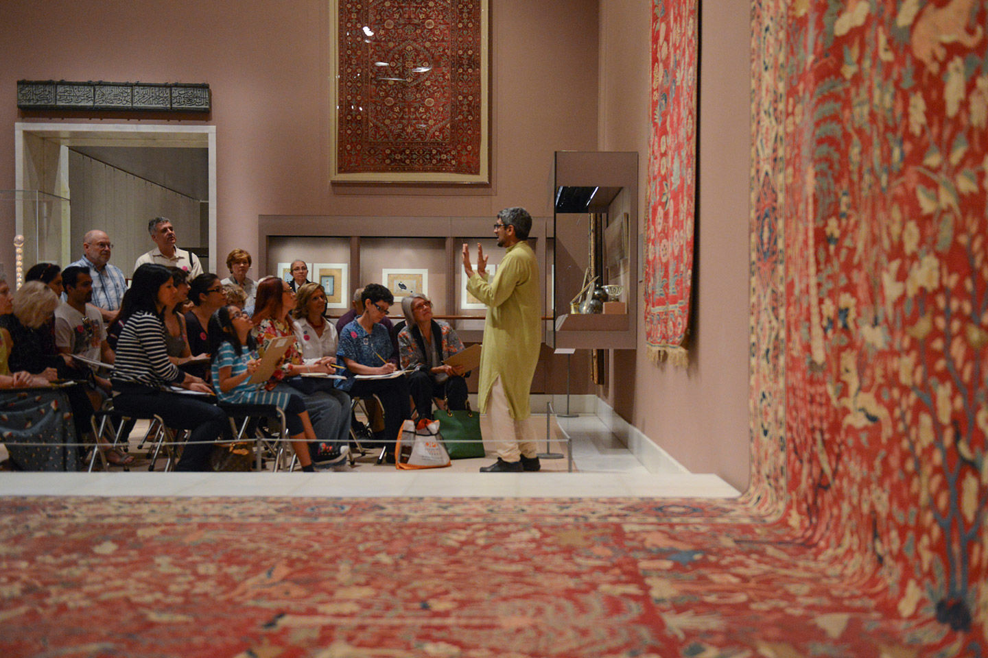 In the near distances a gallery guide talks to a seated crowd within a gallery of hung Islamic carpets